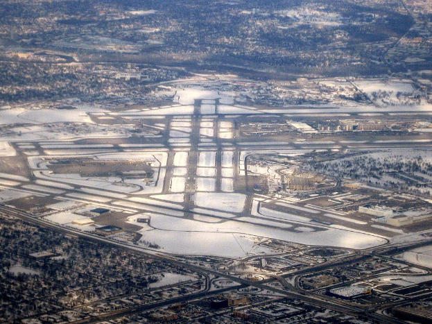 Luftaufnahme eines städtischen Flughafens umgeben von schneebedeckten Straßen, Gebäuden und Bäumen in einer winterlichen Landschaft.