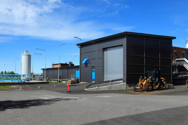 Großes Industriegebäude mit Bulldozer vorne, umgeben von Straßeninfrastruktur und Fahrzeugen, unter einem bewölkten Himmel.