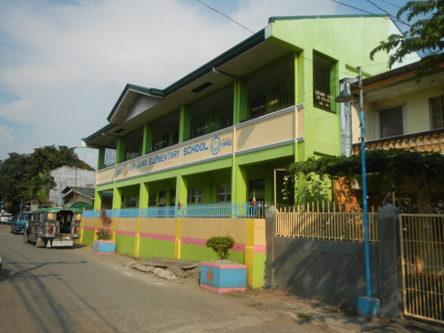 Ein Schulgebäude mit einer grünen und gelben Fassade, das neben einer Straße mit Fahrzeugen, Topfpflanzen, einem Zaun, einem Tor, einem Straßenpfahl, Bäumen, Drähten und einem bewölkten Himmel steht.