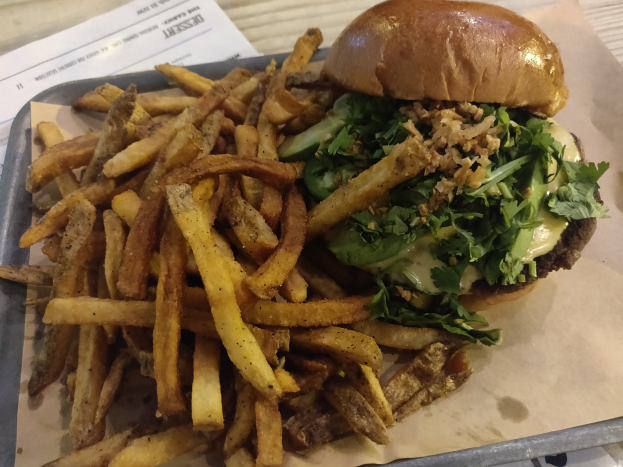 Ein Tablett mit einem Hamburger, der mit Salat, Tomate und Zwiebeln belegt ist, und goldenen, knusprigen Pommes frites, das auf einem Tisch mit einem Blatt daneben steht.