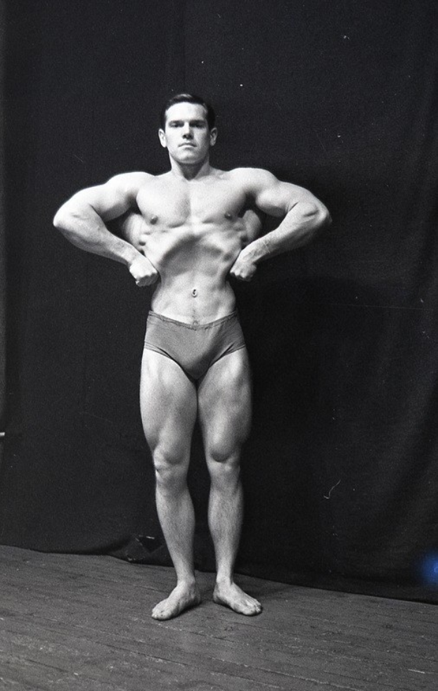 Bodybuilder auf einer Bühne mit Händen in den Hüften stehend, in einem schwarzen und weißen Outfit, vor einem Vorhang.