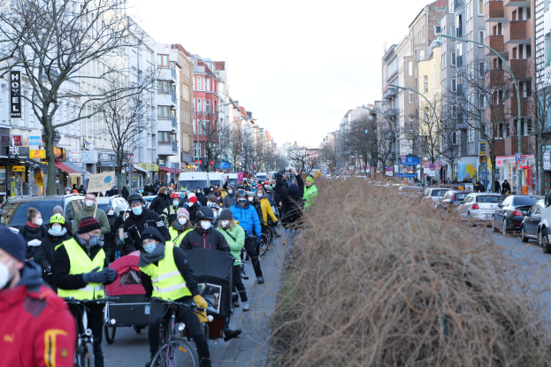 Eine große Gruppe von Menschen in Masken und Sicherheitswesten auf Fahrrädern auf einer von Bäumen gesäumten Straße mit Gebäuden, Laternen, Texttafeln, Fahrzeugen, trockenem Gras und einem klaren blauen Himmel.