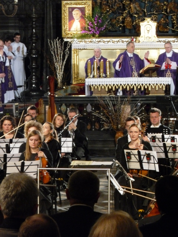 Eine Gruppe von Menschen, die Geigen spielen, mit Noten vor sich, während im Hintergrund drei Kirchenväter hinter einer Bibel stehen, mit einem Foto Rahmen und Verzierungen drumherum.
