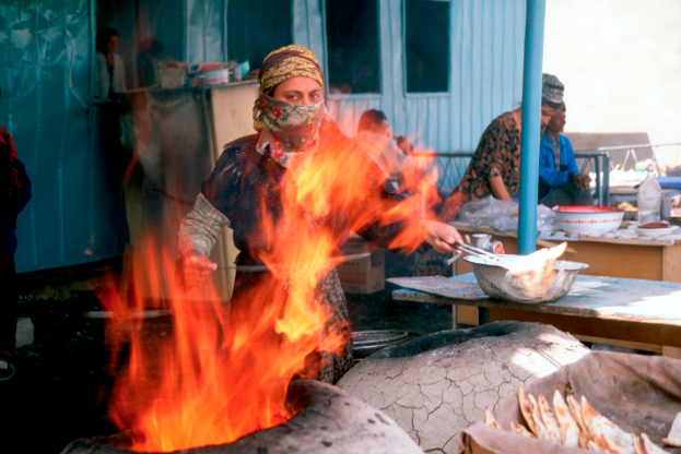 Eine Frau Kocht Essen Auf Einem Feuer Vor Einem Geb├Ąude Mit Fenstern Und Einer T├╝r, W├Ąhrend Menschen Um Einen Tisch Mit Schalen, Flaschen Und Anderen Gegenst├Ąnden Sitzen, Mit Einem Pfahl Und Einer Wand Im Hintergrund.