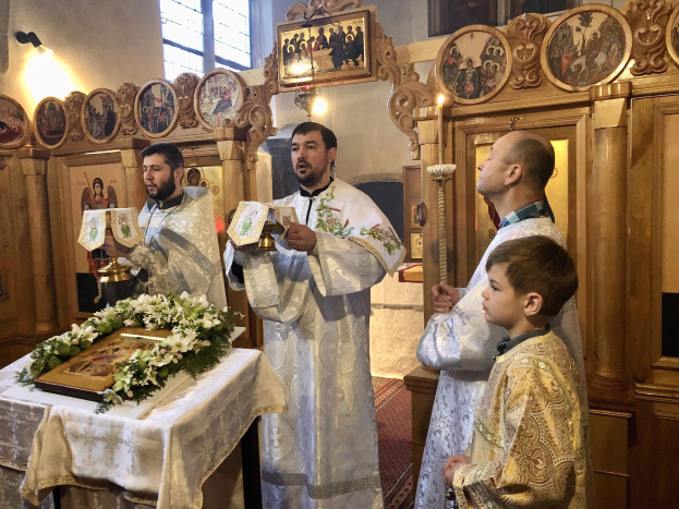 Eine Gruppe von Menschen steht um einen Tisch in einer Kirche, hält Bücher in den Händen, mit einem Tuch und einem Blumenstrauß auf dem Tisch, Fotorahmen, Lampen, einem Fenster und einem hölzernen Objekt im Hintergrund.