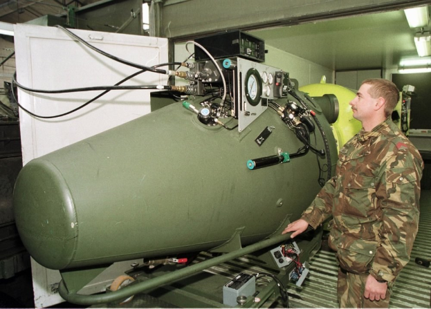 Ein Mann in militärischer Uniform steht neben einer großen grünen Bombe in einer unordentlichen industriellen Umgebung mit Maschinen, Drähten und beleuchtetem Hintergrund, mit Text unten.