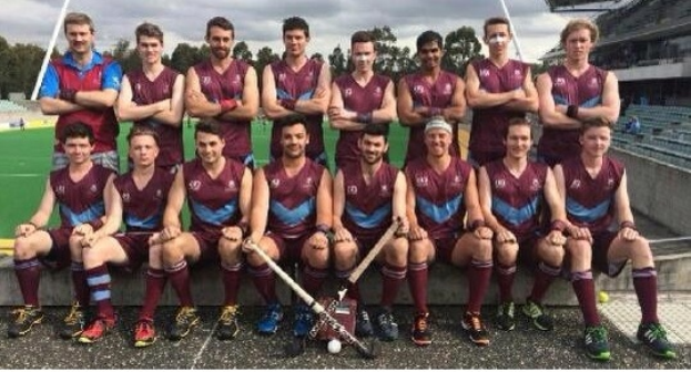 Eine Gruppe von Männern in Sportuniformen steht zusammen auf einem Feld und hält Hockey-Schläger, feiert ihren Sieg mit einem Stadion und Zuschauern im Hintergrund.