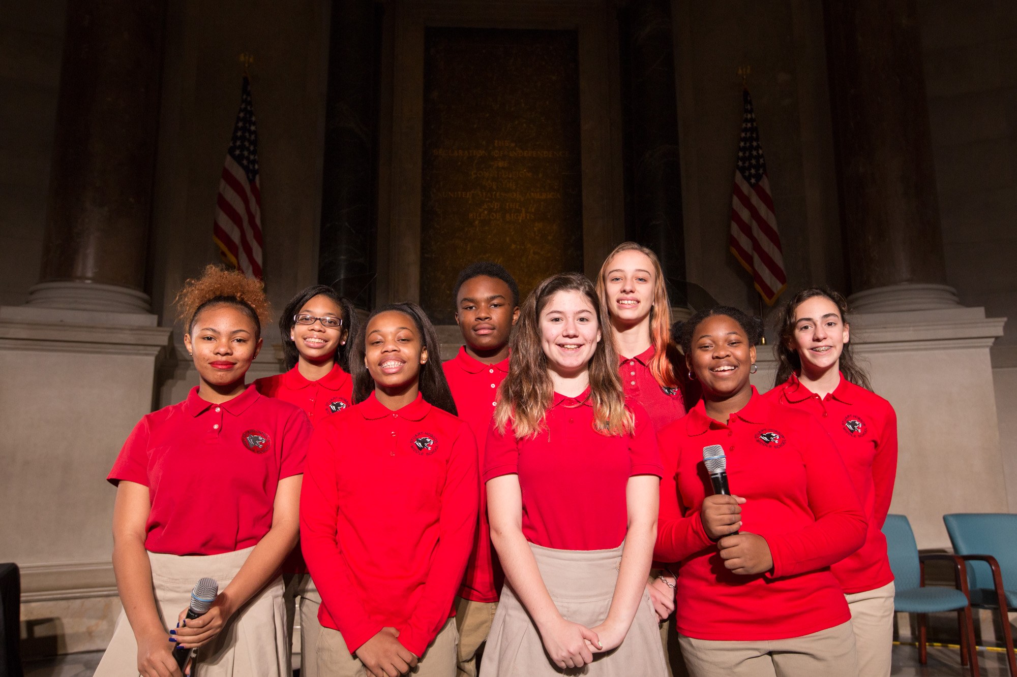 Gruppe junger Frauen in roten Shirts, die strahlend Mikrofone halten und vor Stühlen, Säulen, Fahnen und einer Wand stehen.