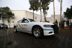 Weißer Polizeiwagen vor einem Gebäude mit einer Gruppe von Menschen in bunten Kleidern und Mützen, Bäumen und einem klaren blauen Himmel in Mexiko-Stadt.