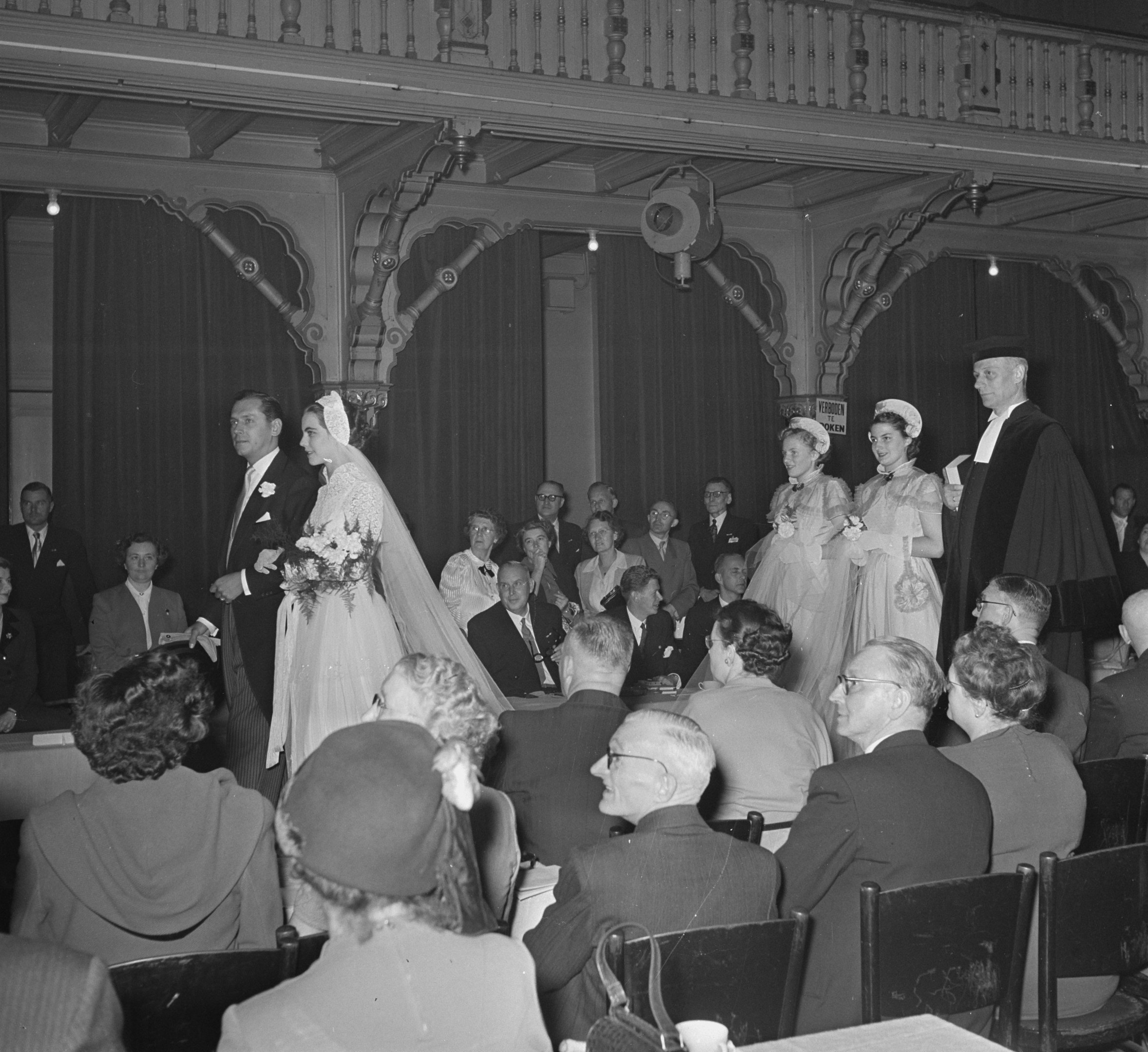 Schwarz-weiß-Foto einer Hochzeitszeremonie in einem großen Raum mit sitzenden und stehenden Gästen, einige halten Blumensträuße, Tische mit Tassen und Gegenständen sowie Vorhänge mit Deckenleuchten im Hintergrund.