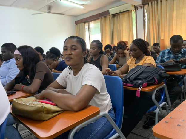 Eine Gruppe von Menschen, die an Tischen in einem classroom an der University of Ghana sitzen, mit Taschen und Gegenständen auf den Tischen, Fenstern mit Vorhängen, einer Klimaanlage an der Wand und Lampen und Ventilatoren an der Decke im Hintergrund.
