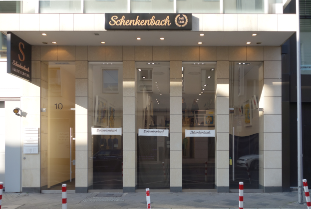 Ein modernes Gebäude in Berlin, Deutschland, mit Glaswänden und -türen, einem Schild mit der Aufschrift "Shenkenbach Haute Couture", einer Überwachungskamera, Papieren an einer Wand, Deckenleuchten, Pfosten und einer Gruppe von Menschen vor dem Gebäude.
