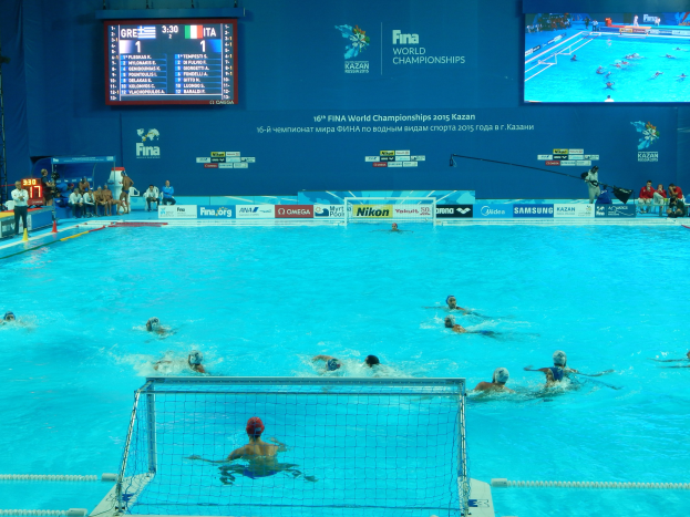 Wasserballspieler bei den 2016 FINA-Weltmeisterschaften in Kazan im Pool antretend, mit Zuschauern auf der linken Seite und einer Anzeigetafel mit Event-Branding im Hintergrund.