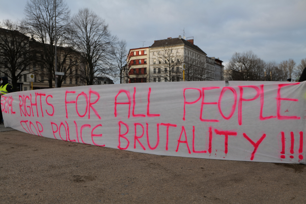 Eine Gruppe von Menschen hält ein Banner mit der Aufschrift 'Rechte für alle Menschen Stoppt Polizeigewalt' auf dem Boden stehend mit einem Straßenschild, einem Schild, Bäumen, Gebäuden mit Fenstern und einem bewölkten Himmel im Hintergrund.