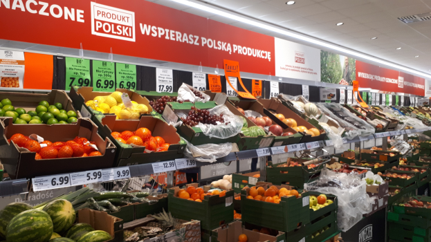 Ein Geschäftsraum mit verschiedenen Früchten und Gemüsen in Kartons mit Preisschildern, Wandtafeln mit Text und Bildern und Deckenbeleuchtung.