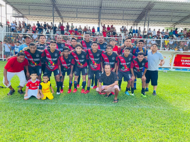 Mannergruppe feiert auf einem Fußballfeld mit einem Siegesschild, auf dem 'Fotos Ugarte U-17 vs U-18' steht, unter einem bewölkten Himmel.