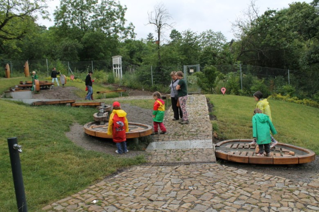 Kinder spielen in einem Park mit grünem Gras, Bäumen und verstreuten Gegenständen, unter einem sichtbaren Himmel, mit einem Weg, der zum Bereich führt, und einem Pfahl auf der linken Seite.