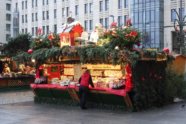 Eine Person steht vor einem Weihnachtsmarktstand in einem Stadtplatz, umgeben von festlichen Dekorationen und anderen Menschen, mit Gebäuden und einem Laternenmast im Hintergrund.