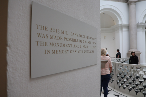 Eine Gruppe von Menschen in einem großen, hellen Innenraum mit einer Tafel an der Wand, einem Zaun, Säulen, Deckenleuchten und einem Schild über die 2013er Millbank-Renovierung, finanziert von den Monument- und Linbury-Stiftungen zum Gedenken an Simon Sainsbury.