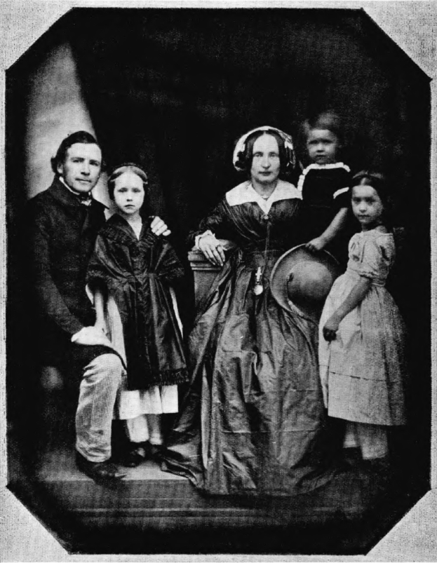 Ein Schwarz-Weiß-Foto einer Familie, die auf einer Treppe posiert, mit einer Frau in einem Kleid, die einen Hut hält und zwei Kinder neben ihr.