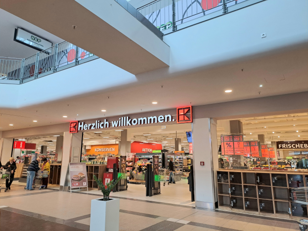 Das Innere eines Einkaufszentrums mit Menschen, die umhergehen, Geschäften, Schildern mit Text, Lampen, Geländern und einem Topf mit einer Pflanze unten; der Text auf den Schildern lautet "Herzlich willkommen".
