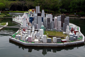 Ein detailliertes Miniaturmodell einer Stadtlandschaft, umgeben von Wasser, mit Gebäuden, Grünflächen, einer Brücke, Menschen und Bäumen vor einem Himmel als Hintergrund.