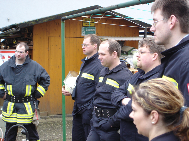 Eine Gruppe von Männern in blauen Feuerwehranzügen, die nach links schauen und vor einem kleinen Holzhäuschen und Rohrrahmen stehen.