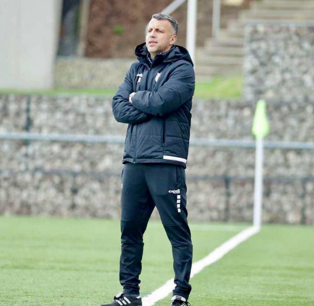 Ein Mann steht auf einem Fußballfeld mit verschränkten Armen und trägt eine schwarze Jacke und Hose, im Hintergrund ist ein Zaun, eine Treppe und eine Wand zu sehen.