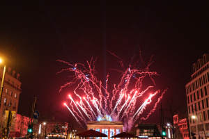Eine belebte Stadtstraße in Berlin am Silvesterabend, voller Menschen, Fahrzeuge und Gebäude, beleuchtet von Lichtern und Feuerwerk am Himmel.
