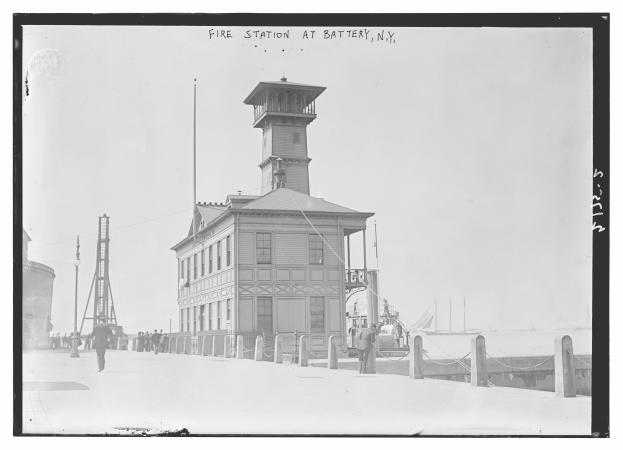 Ein Schwarz-Weiß-Foto einer Feuerwache in Battery, New York, mit einem Gebäude, Menschen, Mästen, einem Zaun, Booten auf dem Wasser und einem bewölktem Himmel.