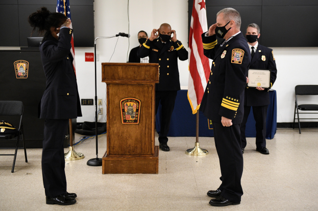 Eine Gruppe uniformierter Personen in Masken steht vor einem Podium mit Mikrofon, umgeben von Flaggen, Stühlen und Tischen, während einer Vereidungszeremonie für einen neuen Feuerchef, wobei eine Person ein Zertifikat hält.