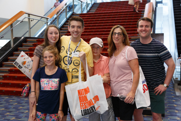 Eine Gruppe lächelnder Menschen mit Taschen steht vor einer Rolltreppe im National Book Festival 2019, mit Treppen, Geländern und einer Wand im Hintergrund.