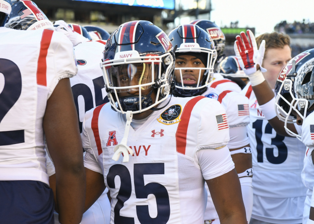 Gruppe von Männern in Football-Uniformen auf einem Feld mit Helmen und Handschuhen, Hintergrund zeigt eine unscharfe Menge, ein Scoreboard mit 'Auburn vs Virginia Tech - 11/28/15 College Football Pick, Odds, and Prediction' und Himmel.