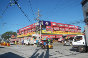 Eine belebte Straße mit einem Lastwagen, mehreren Fahrzeugen, Fußgängern, einem Strommast mit Drähten, Bäumen und einem klaren blauen Himmel im Hintergrund.