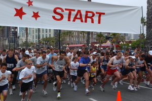 Gruppe von Menschen, die bei einem Marathon laufen, mit einem Verkehrskegel im Vordergrund und einem Banner mit Text im Hintergrund, umgeben von Bäumen, Laternen, Gebäuden und einem klaren blauen Himmel.