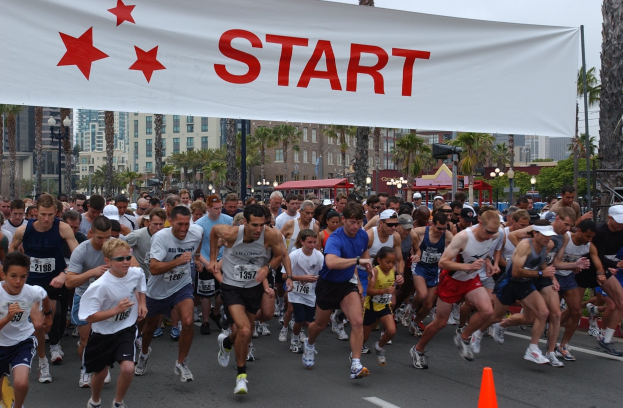 Gruppe von Menschen, die bei einem Marathon laufen, mit einem Verkehrskegel im Vordergrund und einem Banner mit Text im Hintergrund, umgeben von Bäumen, Laternen, Gebäuden und einem klaren blauen Himmel.