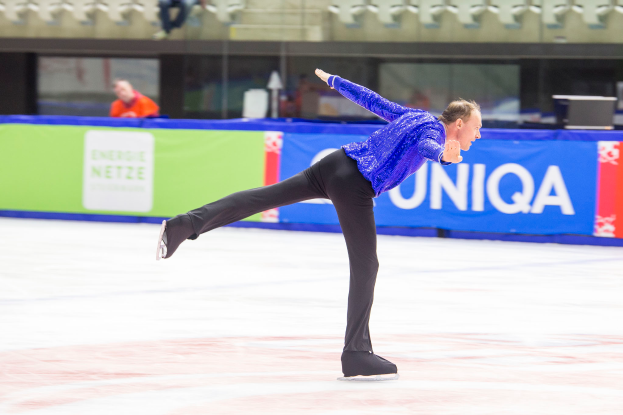 Ein Mann in einem blauen Hemd und schwarzen Hosen fährt auf einer Eisbahn mit einem Banner "World Figure Skating Championships - Day 2" im Hintergrund und einigen Menschen auf den Tribünen.