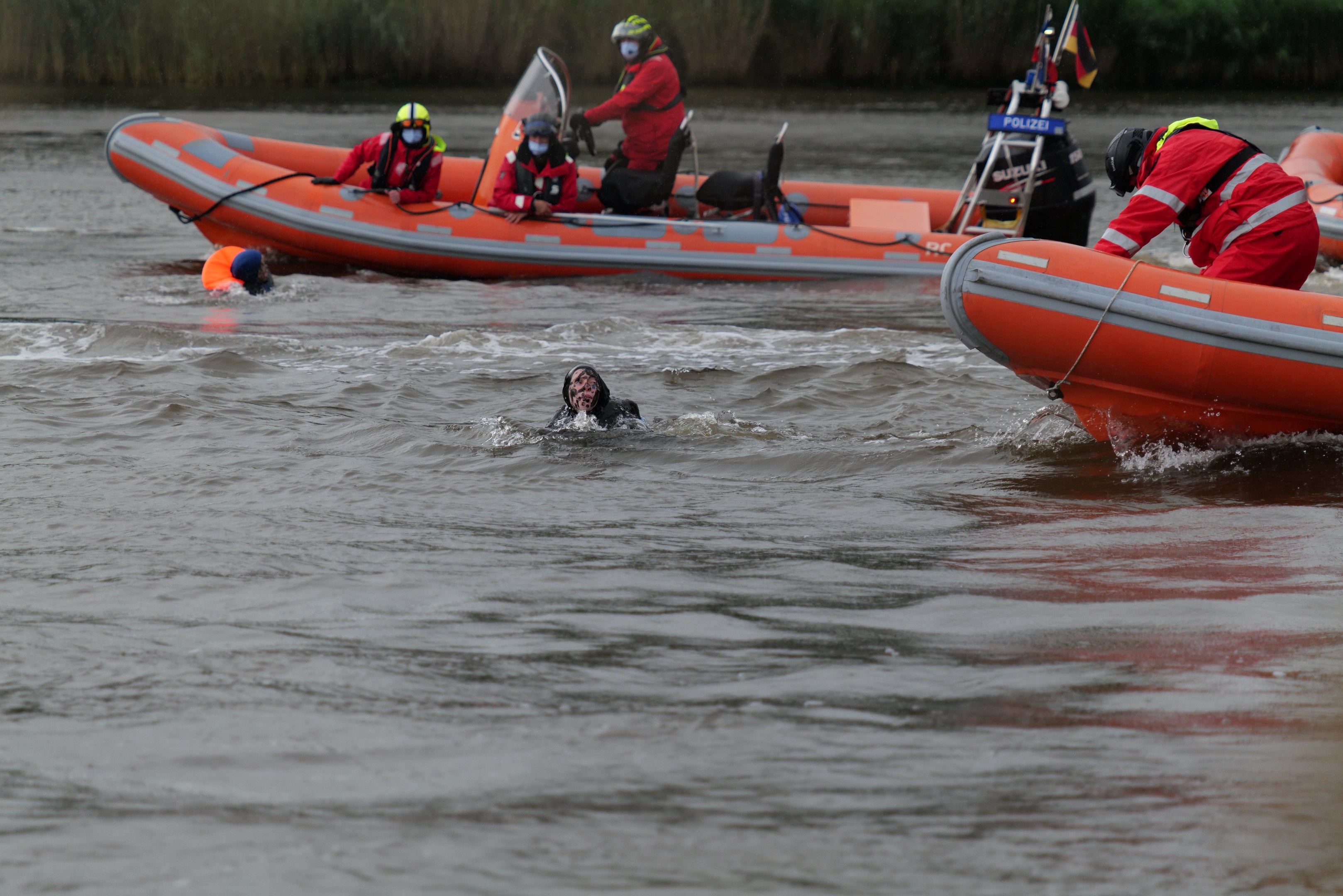 Gruppe von Menschen in einem aufblasbaren Boot auf einem Fluss bei der Hilfe von zwei Personen im Wasser, alle tragen Schwimmwesten und Helme, mit Vegetation im Hintergrund.