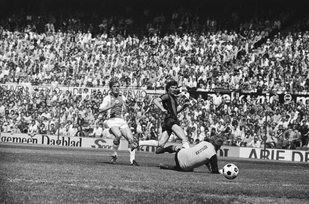 Ein Schwarz-Weiß-Bild einer Gruppe von Männern, die auf einem Feld Fussball spielen, mit Zuschauern im Hintergrund und einer Fahne mit der Aufschrift "Bundesliga 1978-79 Ajax 1-1 Hannover 96".