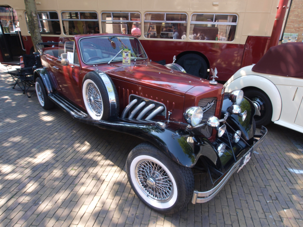Ein Oldtimer neben einem Doppeldeckerbus auf einem Kopfsteinpflaster, mit Stühlen und einem Baum im Vordergrund, einem Gebäude im Hintergrund und Menschen im Bus.