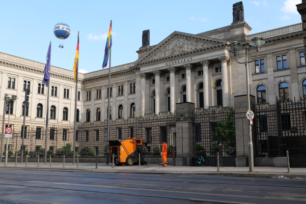 Großes beiges Gebäude mit vielen Fenstern, der Deutsche Bundestag in Berlin, umgeben von Straßenmöbeln, Fahrzeugen, Fußgängern und Bäumen unter einem bewölkten Himmel.