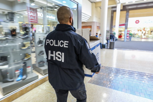 Ein Polizist in einer schwarzen Jacke geht durch ein Einkaufszentrum und trägt eine Kiste, mit Menschen, einem Müllcontainer, Säulen, Deckenlampen und Schuhregalen im Hintergrund.