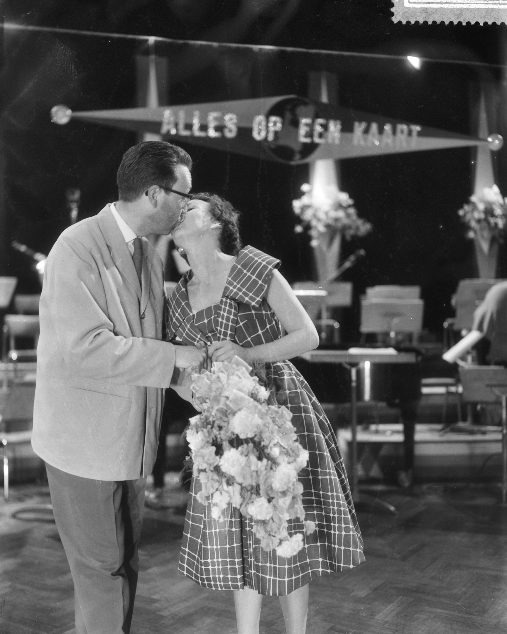 Ein Mann und eine Frau umarmen und küssen sich auf einer Bühne, während die Frau einen Blumenstrauß hält.