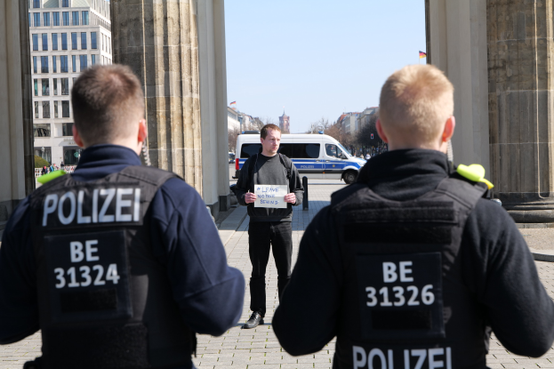 Drei Polizeibeamte stehen vor einem Gebäude, mit einem Mann, der ein Papier zwischen ihnen hält, flankiert von Säulen, während Fahrzeuge, Menschen, Bäume und Gebäude den Hintergrund unter dem Himmel ausfüllen.