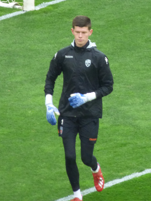 Ein Mann in einer schwarzen Jacke und blauen Handschuhen geht auf einem Fußballfeld mit einem Tor im Hintergrund, Fußballhandschuhe tragend.