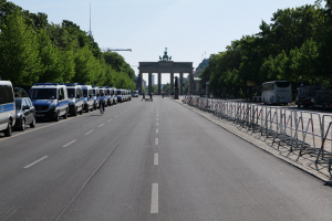 Lange Reihe von Polizeiwagen, die an der Straße vor dem Brandenburgertor geparkt sind, mit Fahrradfahrern und Füüßgängern, Barrieren, Bäumen und einem Bogen mit Statuen im Hintergrund.