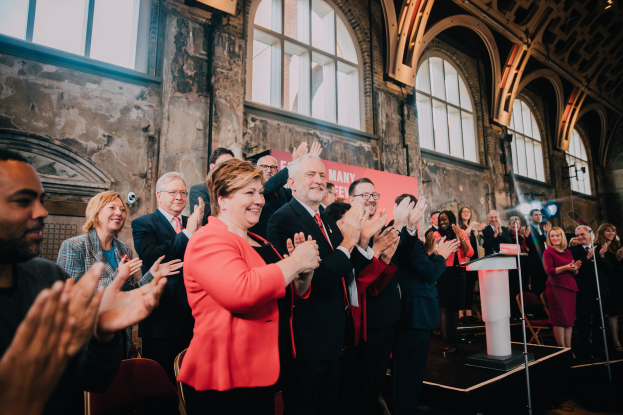Eine Gruppe von Menschen klatscht feierlich vor einem Publikum, mit einem Podium und einem Mikrofon auf der rechten Seite und einem Banner mit der Aufschrift "Covid-19 New York City Mayor Bill de Blasio" im Hintergrund.