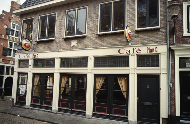 Ein Gebäude mit Fenstern und Türen, ein Schild mit der Aufschrift "Café Plan C in Amsterdam, Niederlande", ein Straßenschild, Bäume und ein bewölkter Himmel.