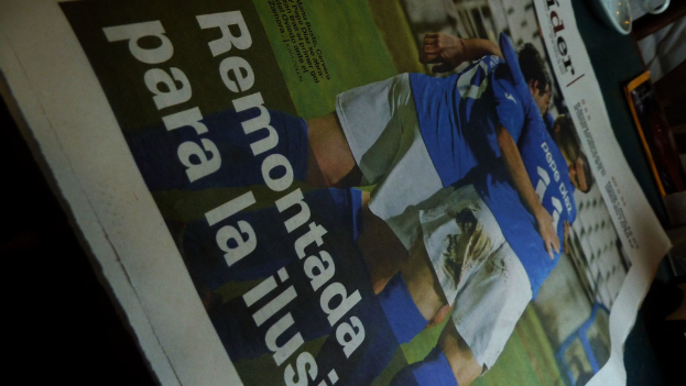 Eine Zeitung auf einem Tisch zeigt drei Männer in blauen Hemden, möglicherweise beim Fußballspielen, mit Text unten im Bild.