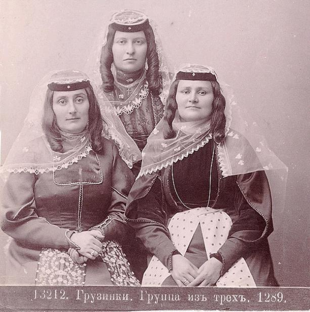 Drei Frauen in traditionellen russischen Hochzeitskleidern mit farbenfroher Stickerei und Perlen stehen vor einer einfarbig weißen Wand in einem Schwarz-Weiß-Foto.